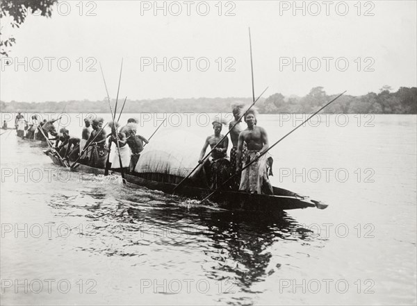 Canoes near Kandahar Island. A group of semi-naked men wearing traditional dress and headgear, paddle canoes past Kandahar Island. Near Livingstone, Northern Rhodesia (Zambia), 12 November 1910., South (Zambia), Zambia, Southern Africa, Africa.