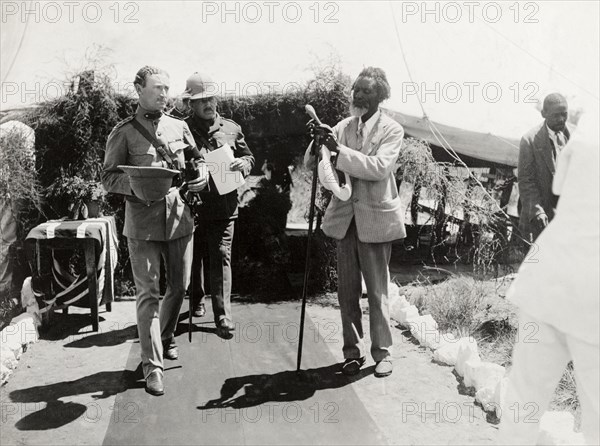 Entrance to a Presentation of Address. An African man, dressed in a Western-style suit, adjusts a long staff at the entrance to a Presentation of Address for the Duke and Duchess of Connaught. Arriving guests file past him along a red carpet. Gwelo, Southern Rhodesia (Gweru, Zimbabwe), 16 November 1910. Gweru, Midlands, Zimbabwe, Southern Africa, Africa.