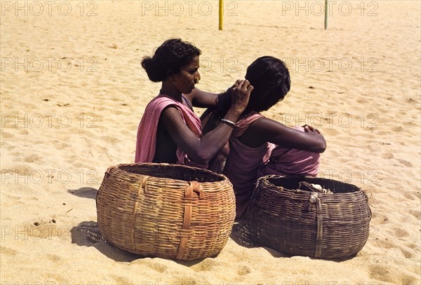 Grooming on an Indian beach. Two women dressed in pink saris sit on a sandy beach beside two large baskets, one grooming the other's hair. India, circa 1976., Jammu and Kashmir, India, Southern Asia, Asia.