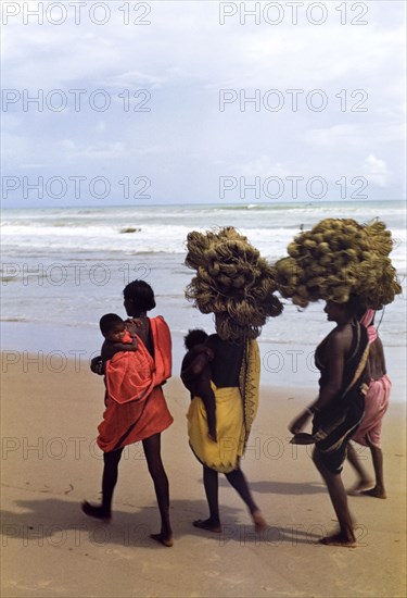 Women carrying bundles of twine. Four women wearing brightly coloured saris walk barefoot along an Indian beach balancing bundles of twine on their heads. Two of the group carry babies under one arm. India, circa 1976., Jammu and Kashmir, India, Southern Asia, Asia.