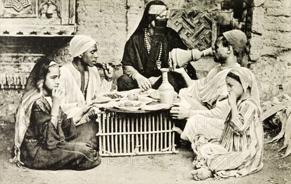 A familiar dinner'. A posed portrait depicting a 'typical' Egyptian meal. A veiled woman adorned with jewellery presides over a generous meal laid out on a large platter. The meal is eaten outdoors from a makeshift table, shared by two men and two girls who sit cross-legged on the ground. Egypt, circa 1925. Egypt, Northern Africa, Africa.