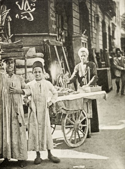 Street peddler, Cairo. A street peddler sells food from a cart whilst two boys in traditional dress look on. Cairo, Egypt, circa 1910. Cairo, Cairo, Egypt, Northern Africa, Africa.