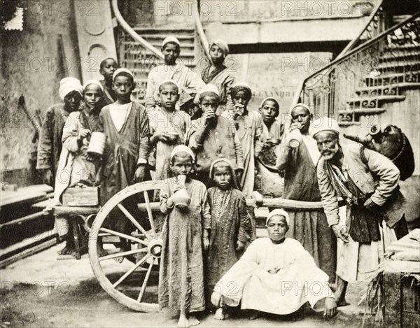 Arab children collect water. Portrait of a group of traditionally dressed Arab children, several of whom hold drinking vessels. The presence of an elderly man, who carries a large jug on his back, suggests that they have probably congregated to collect water. Cairo, Egypt, circa 1910. Cairo, Cairo, Egypt, Northern Africa, Africa.