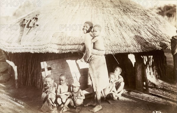 A Matabele family. A Matabele (Ndebele) woman turns to smile for the camera, a young child clinging to her back and five more sitting around her feet. An original caption comments: 'One man may have a doz(en) wives, hence the number of piccininas (children)'. Probably Southern Rhodesia (Zimbabwe), circa 1910. Zimbabwe, Southern Africa, Africa.