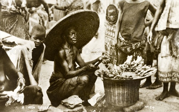 Fish seller, Gold Coast. An elderly woman dressed in a huge, wide brimmed hat, sells fish to passers by on a crowded street. Gold Coast (Ghana), circa 1910. Ghana, Western Africa, Africa.