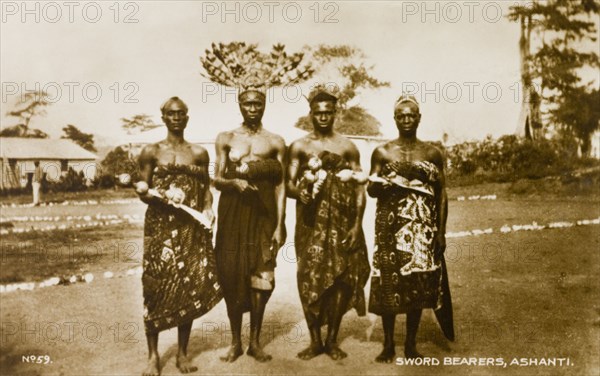 Asante swordbearers. Group portrait of four traditionally dressed Asante (Ashanti) swordbearers. One wears a feathered headdress and each holds a ceremonial sword. Gold Coast (Ghana), circa 1910. Ghana, Western Africa, Africa.