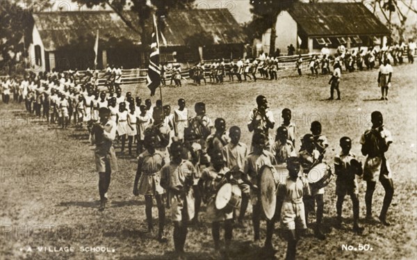 Gold Coast school parade. A procession of uniformed school children parade outdoors, displaying a union jack flag. The boys in front provide a musical accompaniment, playing flutes and drums as they walk. Gold Coast (Ghana), circa 1910. Ghana, Western Africa, Africa.