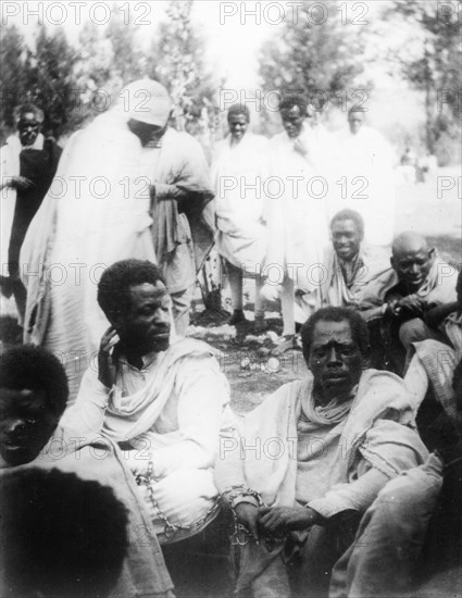 East African slave court. A group of east African slaves gather for the hearing of a slave court, the two nearest men chained together by the wrists. Eastern Africa, circa 1933., Eastern Africa, Africa.