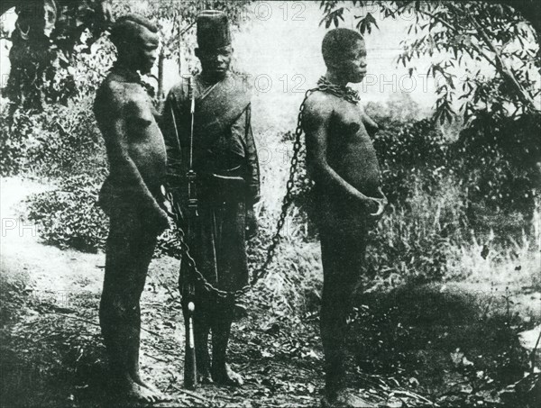 Congo Free State hostages. Two semi-naked women are chained together by the neck under armed guard. They were taken hostage by sentries of the Anglo-Belgian India Rubber company (ABIR) after their husbands fled into the forest to escape slavery. Congo Free State (Democratic Republic of Congo), circa 1905. Congo, Democratic Republic of, Central Africa, Africa.