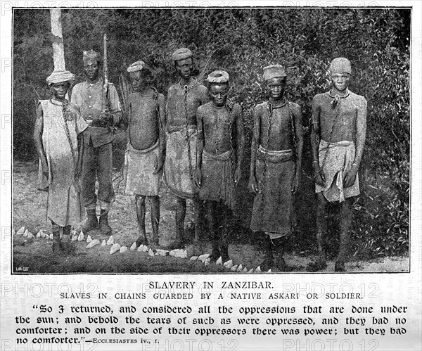 Slaves in Zanzibar. Portrait of a six male slaves, chained together by the neck and guarded by a uniformed soldier. Zanzibar (Tanzania), 1896., Zanzibar Central/South, Tanzania, Eastern Africa, Africa.