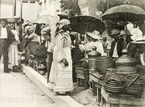 Selling jackass rope tobacco. A line of street traders sit beneath umbrellas as they sell rolls of jackass rope tobacco by the roadside. The term 'jackass rope tobacco' is used in Jamaican colloquial dialect to describe homegrown tobacco that has been twisted into a rope. Kingston, Jamaica, circa 1905. Kingston, Kingston, Jamaica, Caribbean, North America .