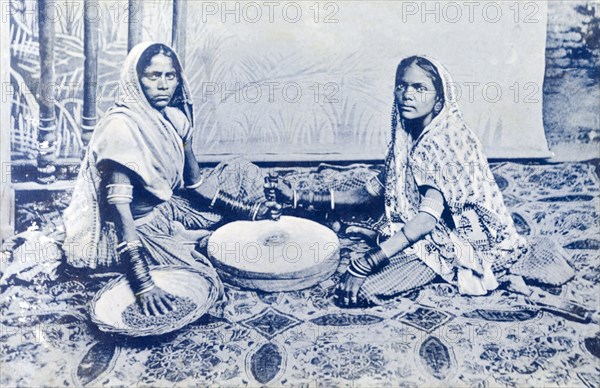 Indian women grinding corn. A posed portrait of two Indian women grinding corn between a pair of millstones. Traditionally dressed in saris and adorned with jewellery, they sit on an oriental rug in front of an artificial backdrop depicting a jungle scene. India, circa 1910. India, Southern Asia, Asia.