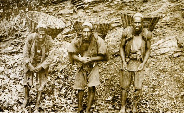 Bheestis' or water carriers. Portrait of three 'bheestis' or water carriers, transporting baskets of bottled water on their backs. The rocky landscape and appearance of the men suggests that the photograph was taken on the Afghanistan-Pakistan border. Probably India (Pakistan), circa 1915. Pakistan, Southern Asia, Asia.