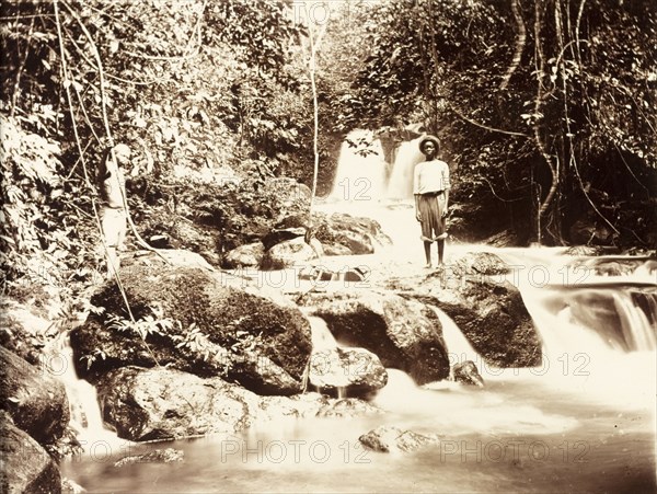 Jungle river in Bioko. Two young men stand on a rock formation straddling a jungle river. The slow shutter speed used by the photographer gives the water an ethereal quality as it cascades over the rocks in waterfalls. Bioko, Equatorial Guinea, circa 1930., Bioko Norte, Equatorial Guinea, Central Africa, Africa.