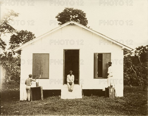 Painting the shutters. Two men paint shutters on the exterior of a small building with a pitched roof, possibly a missionary station, whilst another rests in the doorway. Bioko, Equatorial Guinea, circa 1930., Bioko Norte, Equatorial Guinea, Central Africa, Africa.