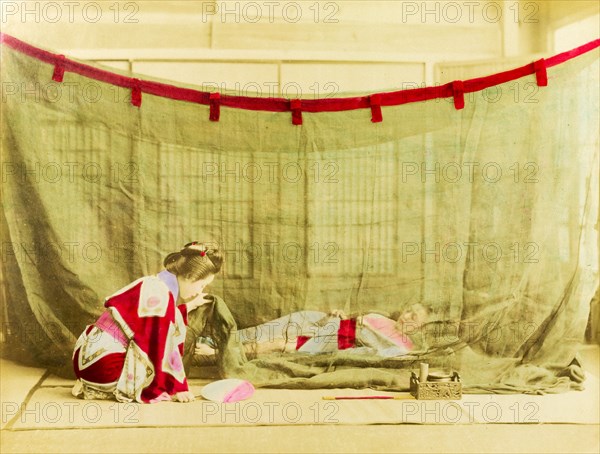 Japanese sleeping arrangements. A kneeling Japanese woman dressed in a kimono lifts the corner of a mosquito net, behind which another woman lies sleeping on a futon. Japan, circa 1910. Japan, Eastern Asia, Asia.