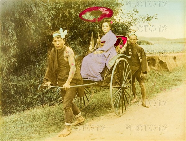 A Japanese rickshaw. A young Japanese woman travels along a rural road in an open rickshaw pulled by a runner. Finely dressed, she wears a traditional kimono and holds a parasol. Japan, circa 1910. Japan, Eastern Asia, Asia.