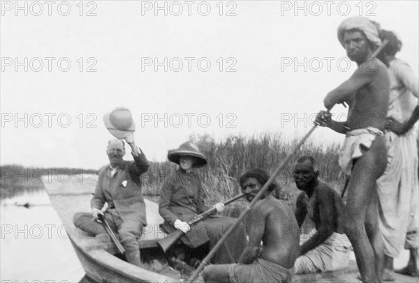 A duck hunt at Christmas. Sir Henry Staveley Lawrence, Collector of Karachi, raises his solatopi hat to the camera during a duck hunt in the countryside. He sits in a canoe piloted by Indian 'shikaris' (professional hunters), in the company of Miss Arthur, who holds a rifle across her knee. Sind, India (Sindh, Pakistan), December 1911., Sindh, Pakistan, Southern Asia, Asia.