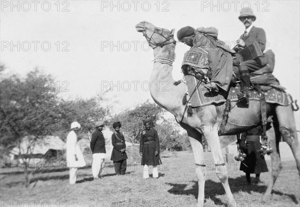 Mr Haywood rides a camel. A European man identified as Mr Haywood sits on a camel behind a 'shikari' (professional hunter), ready to embark on a hunting expedition to the countryside. Probably Sind, India (Sindh, Pakistan), December 1911., Sindh, Pakistan, Southern Asia, Asia.