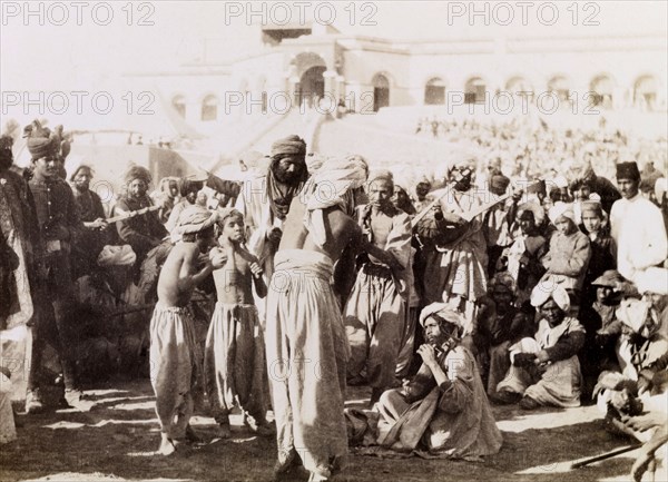 Indian musicians in Sukkur. Indian musicians with stringed instruments play to a gathered crowd, while a man in traditional wrestlers costume and two boys confer with the group. Sukkur, Sind, India (Sindh, Pakistan), circa 1908. Sukkur, Sindh, Pakistan, Southern Asia, Asia.