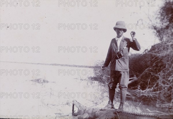 Crocodile hunting in India. Sir Henry Staveley Lawrence, Collector of Karachi, stands on the carcass of a crocodile, which has had its mouth propped open for the camera. He poses beside the Indus River where the animal was shot, wearing a solatopi hat and breeches with a pair of binoculars slung around his neck. Probably Sind, India (Sindh, Pakistan), December 1911., Sindh, Pakistan, Southern Asia, Asia.