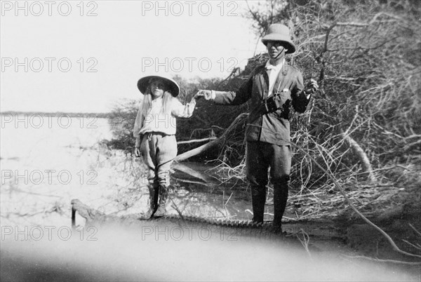 Father and daughter on a crocodile hunt. Sir Henry Staveley Lawrence, Collector of Karachi, holds hands with his daughter Margaret (b.1904), as they stand on the carcass of a crocodile, which has had its mouth propped open for the camera. They pose beside the Indus River where the animal was shot, wearing solatopi hats and breeches. Probably Sind, India (Sindh, Pakistan), December 1911., Sindh, Pakistan, Southern Asia, Asia.