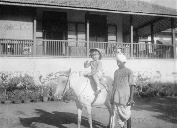 Adam rides a pony, Bombay. A British boy called Adam waves to the camera as he rides a pony in the garden of a colonial house, led by an Indian servant. Bombay (Mumbai), India, 1940. Mumbai, Maharashtra, India, Southern Asia, Asia.
