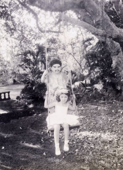 Children on a swing, India. Two British children play on a garden swing. Both wear fancy dress: the girl dressed in a white tutu with flowers in her hair, and the boy dressed in an oriental-style outfit, complete with false moustache. India, circa 1940. India, Southern Asia, Asia.