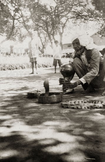 An Indian snake charmer at work. An Indian snake charmer plays a traditional bamboo 'pungi' as he charms a cobra coiled up in a basket in front of him. The striped body of a large python can be seen to the right. Hiwan, Mandi, Simla Hill State (Himachal Pradesh), India, circa 1926. Mandi, Himachal Pradesh, India, Southern Asia, Asia.