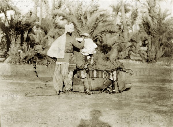 Margaret Lawrence prepares to ride a camel. Margaret Lawrence (b.1904), the daughter of Phyllis and Sir Henry Staveley Lawrence (Collector of Karachi), clambers up onto the back of a saddled camel with the aid of an Indian servant. Karachi, India (Pakistan), circa 1910. Karachi, Sindh, Pakistan, Southern Asia, Asia.