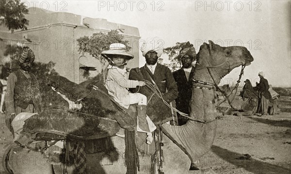 Margaret Lawrence rides a camel. Margaret Lawrence (b.1904), the daughter of Phyllis and Sir Henry Staveley Lawrence (Collector of Karachi), rides a saddled camel under the watchful eye of three Indian servants. Karachi, Sind, India (Sindh, Pakistan), circa 1910. Karachi, Sindh, Pakistan, Southern Asia, Asia.