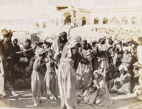 Musicians perform in Sukkur. Spectators at an outdoor event gather to listen to a group of musicians who play a variety of stringed instruments. Sukkur, Sind, India (Sindh, Pakistan), circa 1908. Sukkur, Sindh, Pakistan, Southern Asia, Asia.