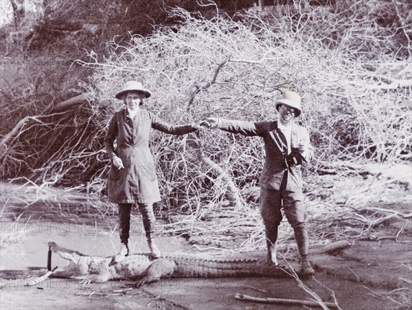 Husband and wife on a crocodile hunt. Sir Henry Staveley Lawrence, Collector of Karachi, holds hands with his first wife Phyllis, as they stand on the carcass of a crocodile, which has had its mouth propped open for the camera. They pose beside the Indus River where the animal was shot, wearing solatopi hats and breeches. Probably Sind, India (Sindh, Pakistan), December 1911. Pakistan, Southern Asia, Asia.