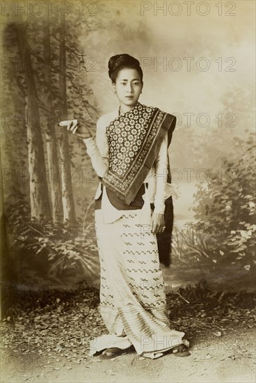 Portrait of a Burmese lady. A posed studio portrait of a young Burmese woman, traditionally dressed in a patterned silk 'longyi' or wraparound skirt. Her hair is worn up and she stands against a back cloth painted with a forest scene. Leaves are scattered around her feet and she holds a 'cheroot' (a type of cigar) in one hand. Sagaing, Burma (Myanmar), circa 1891., Sagaing, Burma (Myanmar), South East Asia, Asia.