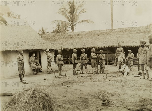British and Indian soldiers in Burma (Myanmar). Armed British and Indian soldiers, possibly a mix of British Army and Military Police officers, mill about outside two thatched roof dwellings. A series of wooden stakes hammered into the ground appear to be fitted with restraints: perhaps a method of detaining captured prisoners. Sagaing, Burma (Myanmar), 1891., Sagaing, Burma (Myanmar), South East Asia, Asia.