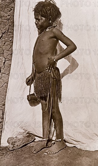 Portrait of a young girl in Omdurman. Professional portrait of a young Sudanese girl, standing barefoot against the backdrop of a white sheet with one hand on her hip. She is semi-naked, wearing only a grass skirt and jewellery, including anklets, bracelets and necklaces. Omdurman, Sudan, circa 1910. Omdurman, Khartoum, Sudan, Eastern Africa, Africa.