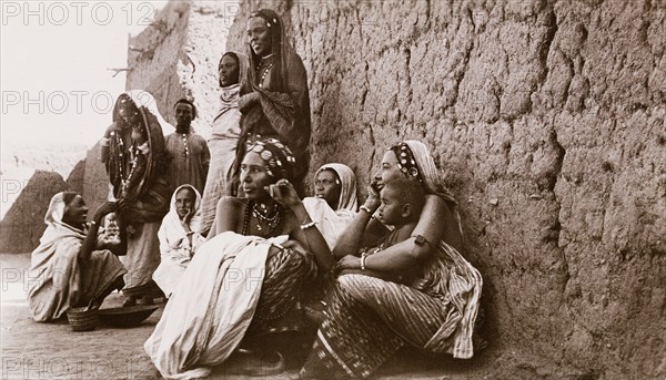 Sudanese women in Omdurman. A group of Sudanese women crouch and stand together beside a high mud wall. They wear traditional dress including decorated headscarves, patterned robes and ornate, beaded jewellery. Omdurman, Sudan, circa 1910. Omdurman, Khartoum, Sudan, Eastern Africa, Africa.