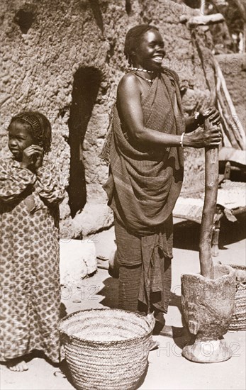 Grinding 'durra', Sudan. A Sudanese woman in traditional dress smiles as she uses a large pestle and mortar to grind 'durra', the grain of a cereal grass that is used to make bread. Omdurman, Sudan circa 1910. Omdurman, Khartoum, Sudan, Eastern Africa, Africa.