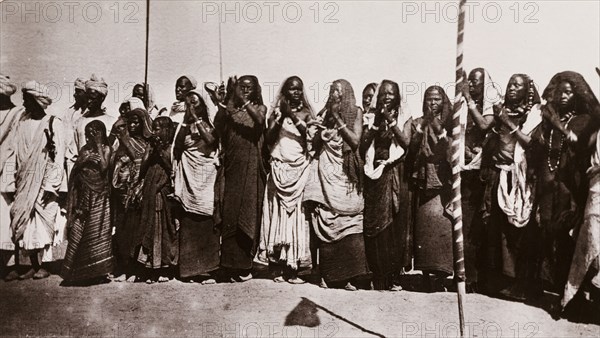 Baggara women clap their hands. A group of Baggara women stand in a line, clapping their hands in unison. They wear traditional dress including long headscarves and beaded necklaces. Geizira (Al Jazirah State), Sudan, circa 1910., Al Jazirah, Sudan, Eastern Africa, Africa.