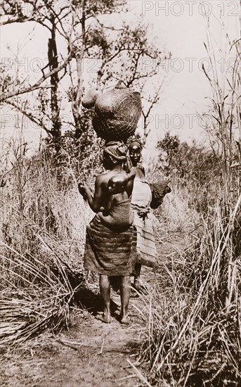 How women travel in the Sudan'. Two Sudanese women in traditional dress walk along an overgrown path. One carries a child on her back, the other balances a bundled load on her head. Sudan, circa 1910. Sudan, Eastern Africa, Africa.