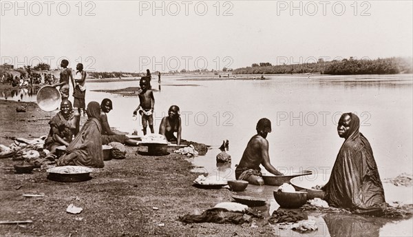 Washing clothes at the river bank, Sudan. Sudanese women and children wash their clothes on the banks of a river, using large, shallow bowls filled with river water. Waw, Sudan, circa 1910. Waw, West Bahr el Ghazal, Sudan, Eastern Africa, Africa.