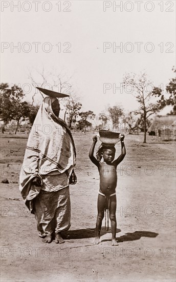 Mother and child carrying bowls. A mother and her child carry bowls on their heads along a village road. Bahr-el-Ghazal region, Sudan, circa 1910., West Bahr el Ghazal, Sudan, Eastern Africa, Africa.