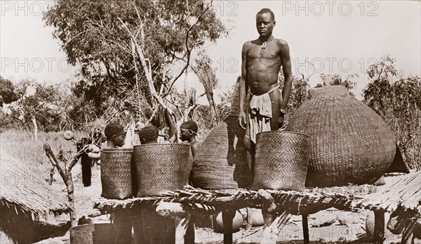 A village granary, Sudan. A man in a loincloth stands on a raised platform between several baskets at a village granary. Three women chat behind him at ground level. Bahr-el-Ghazal region, Sudan, circa 1910., West Bahr el Ghazal, Sudan, Eastern Africa, Africa.