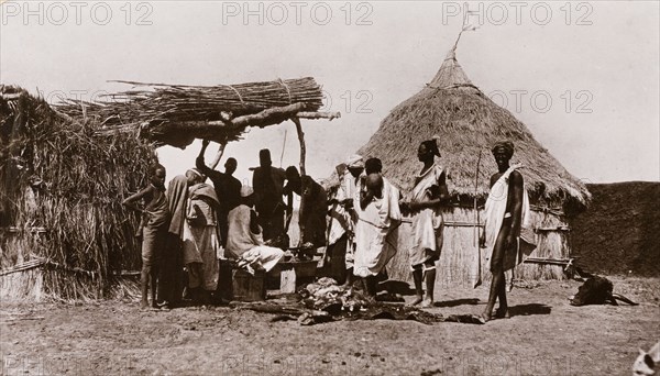 Butcher's shop in a Sudanese village. A line of men queue up outside a village butcher's stall, waiting to be served by an assistant who sits at a workbench beneath a canopy, surrounded by cuts of meat. Upper Nile State, Sudan, circa 1910., Upper Nile, Sudan, Eastern Africa, Africa.
