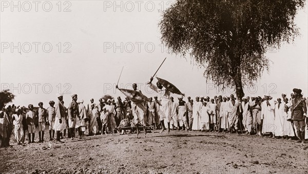 Sudanese men perform a sword display. A crowd of onlookers watch as two Sudanese men perform a sword display. They jump energetically into the air, wielding their swords above their heads with their shields raised. Blue Nile State, Sudan, circa 1910., Blue Nile, Sudan, Eastern Africa, Africa.