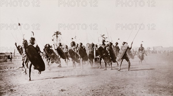 Shilluk warriors charge. A group of Shilluk warriors charge along a dusty plain carrying shields and wielding spears. Upper Nile State, Sudan, circa 1910., Upper Nile, Sudan, Eastern Africa, Africa.