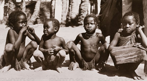 Four Sudanese children. Four semi-naked Sudanese children squat on the ground in a line. Their hair is braided and one wears a beaded necklace. Khartoum, Sudan, circa 1910. Khartoum, Khartoum, Sudan, Eastern Africa, Africa.