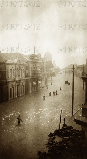 Flooded street in Karachi. A small band of people wades towards a partially submerged vehicle along a flooded street in Karachi. The water is knee-deep, almost reaching the ground-floor windows of the surrounding buildings. Karachi, India (Pakistan), circa 1910. Karachi, Sindh, Pakistan, Southern Asia, Asia.