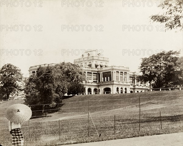 Government House, Singapore. Government House stands on a rise in landscaped gardens behind a wire fence. A person in local dress holding an Asian umbrella stands with their back to the camera in the left foreground. Singapore, Straits Settlements (Singapore), circa 1895. Singapore, Central (Singapore), Singapore, South East Asia, Asia.