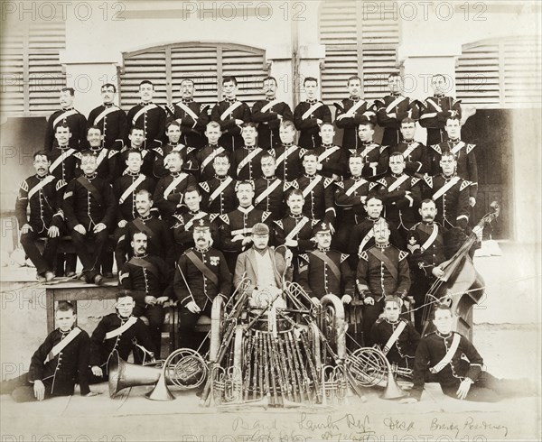 British Army military band with instruments. A British Army military band pose for a group portrait, seated in rows behind their musical instruments, which include clarinets, brass horns and a doube bass. The central figure wears a casual jacket and cap and holds what appears to be a squash racquet. Southern Asia, circa 1895., Southern Asia, Asia.